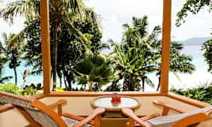 Tremønert terrasse med to stoler og et bord med utsikt over tropiske palmer og blått hav. (AI-generert tekst)