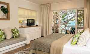 Bilde av et hotellrom med en komfortabel dobbeltseng og en sittegruppe, en TV på et hvitt møbel og utsikt mot havet fra en møblert balkong. (AI-generert tekst)