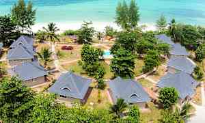 Flyfoto av bolighus med hager og gangveier ved en strand med turkis hav. (AI-generert tekst)