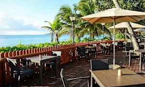 Uteservering ved sjøen med bord, stoler og parasoller, omgitt av grønne planter og palmetrær. (AI-generert tekst)