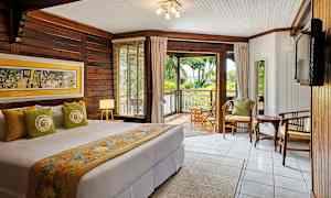 Soverom med dobbeltseng, dekorert med fargerike tekstiler og veggkunst, har trepanelvegger og et stort vindu med utsikt over en frodig veranda. (AI-generert tekst)