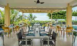 En utendørs restaurant med bord og stoler omgitt av grøntområder og utsikt over havet. (AI-generert tekst)