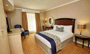 Et hotellrom med en oppredd seng, en lenestol, et skrivebord og et rundt speil på veggen. (AI-generert tekst)