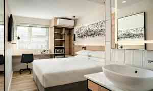 Et moderne hotellrom med en dobbeltseng, et skrivebord med stol, en veggmontert TV, samt en åpen garderobe og en liten vask med speil. (AI-generert tekst)