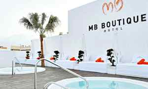 Et hotellområde med hvite solsenger og oransje håndklær ved et basseng, samt en palme og hvite vegger. (AI-generert tekst)