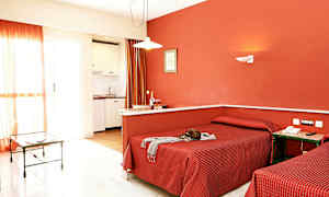 Et hotellrom med to enkeltsenger, røde sengetepper og en rød vegg, samt et lite kjøkken og et glassbord i bakgrunnen. (AI-generert tekst)