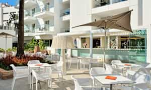 Et utendørsområde med bord, stoler og parasoller foran en bygning. (AI-generert tekst)