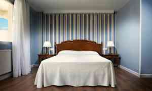 Hotellrom med seng i midten, to nattbord med lamper og stripete blå tapet. (AI-generert tekst)