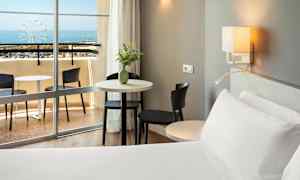 Et hotellrom med en seng, bord og stoler nær en balkong med utsikt over havet. (AI-generert tekst)