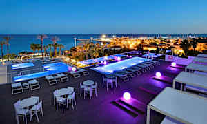 Utendørs bassengområde med utsikt over havet og en småbåthavn, belysning i mørket. (AI-generert tekst)