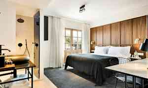 Et hotellrom med en stor seng med mørkgrå sengeteppe, en trepanel som sengegavl, et arbeidsområde med bord og stol, samt et synlig bad med dusj bak en glassdør. (AI-generert tekst)