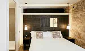 Et hotellrom med en dobbeltseng, to hengende lamper og et innebygd lysfelt på treveggen bak sengen. (AI-generert tekst)