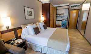 Et hotellrom med en stor seng, puter, nattbord og en liten kjøkkenkrok med skap. (AI-generert tekst)