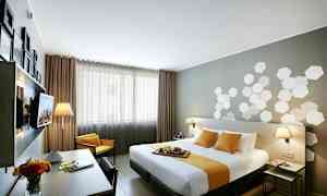 Et moderne hotellrom med en dobbeltseng dekket av hvitt og gult sengetøy. I rommet er det et skrivebord med en blomsterkanne og lamper på nattbordene. (AI-generert tekst)