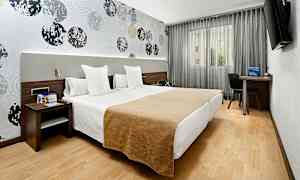 Et hotellrom med to enkeltsenger plassert ved siden av hverandre. Veggene er dekorert med et mønster av sirkler, og det finnes en TV, skrivebord og stol ved vinduet. (AI-generert tekst)