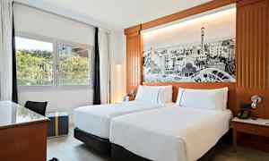 Et hotellrom med to enkeltsenger, et skrivebord, utsikt mot grøntområder gjennom vinduet, og en vegg med et stort svart-hvitt bilde som dekor. (AI-generert tekst)