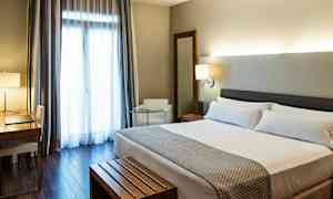 Et hotellrom med en dobbeltseng, et skrivebord med stol, en lenestol og et stort vindu med gardiner. (AI-generert tekst)