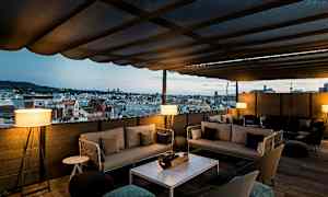 En terrasse med tak og utsikt over en bys silhuett. Terrassen er møblert med sofaer og bord. (AI-generert tekst)