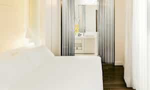 Et hotellrom med en hvit seng og et moderne bad synlig gjennom vertikale svarte paneler. (AI-generert tekst)