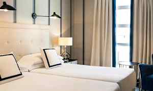 To enkeltsenger står ved siden av hverandre med puter og hvitt sengetøy. Bak sengene er det en lys vegg med stripemønster. Ved sengen står et bord med en bordlampe, og et vindu med gardiner vises i bakgrunnen. (AI-generert tekst)