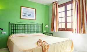 Et soverom med en grønn vegg, en seng med beige sengeteppe, en svart sengegavl, og et vindu med stripete gardiner. (AI-generert tekst)