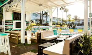 En utendørs lounge under en hvit pergola med møbler og planter, med utsikt over et basseng og omkringliggende bygninger. (AI-generert tekst)