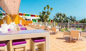 En utendørsterrasse med bord og stoler, omgitt av palmer og fargerike bygninger. (AI-generert tekst)