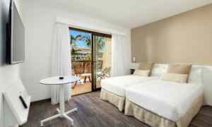 Et moderne hotellrom med to enkeltsenger, et lite bord og en veggmontert TV. Rommet har lyse farger og et vindu som leder ut til en balkong med utsikt mot palmer. (AI-generert tekst)