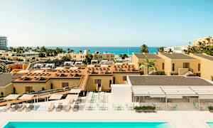 Et bilde av en ferieby med gule bygninger, et basseng, solsenger og havutsikt i det fjerne. (AI-generert tekst)