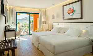 Et lyst og moderne hotellrom med to senger, en sittegruppe og et skrivebord. En balkong med utsikt over landskapet er synlig i bakgrunnen. (AI-generert tekst)