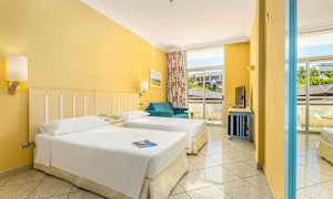 Et hotellrom med to enkeltsenger, en blå sofa og store vinduer med utsikt utenfor. (AI-generert tekst)