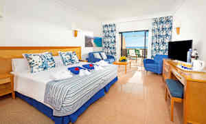 Et hotellrom med en dobbeltseng, sittegruppe og uteplass. Utsikten viser havet. (AI-generert tekst)