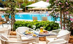 Et bord med mat og drikke står på en uteservering nær et basseng omgitt av blomstrende planter og parasoller. (AI-generert tekst)