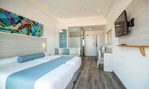 Et moderne hotellrom med en dobbeltseng dekorert med blå tekstiler, en sitteplass med puter, et skrivebord og en veggmontert TV. (AI-generert tekst)