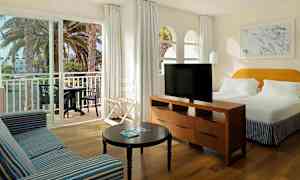 Bilde av et hotellrom med en stor seng, en sofa, et bord og en TV. En balkong med bord og stoler er synlig gjennom skyvedører. (AI-generert tekst)