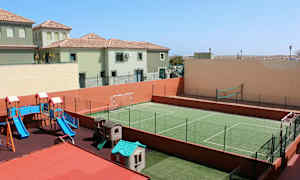 Bildet viser en utendørs lekeplass med sklie og lekestue samt en grønn inngjerdet sportsbane for fotball og volleyball. I bakgrunnen sees bolighus. (AI-generert tekst)
