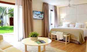 Et lyst hotellrom med en stor seng, veggmontert TV og en sittegruppe foran et rundt bord. Store gardiner med utsikt mot en hage. (AI-generert tekst)