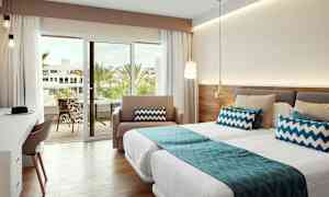 Et godt opplyst hotellrom med senger, blå dekorative puter og en glassdør som fører til en balkong med utsikt over palmer. (AI-generert tekst)