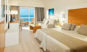 Et hotellrom med dobbeltseng, sittegruppe og balkong med havutsikt. (AI-generert tekst)