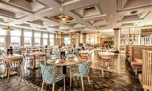 Et elegant restaurantområde med runde bord og mønstrede stoler, opplyst av taklamper og naturlig lys gjennom store vinduer. (AI-generert tekst)