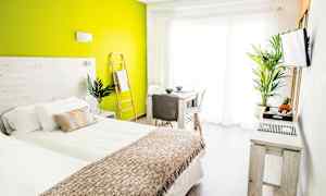 Et lyst og moderne soverom med grønn aksentvegg, seng med hvit og beige sengetøy, samt bord og stoler nær et stort vindu. (AI-generert tekst)