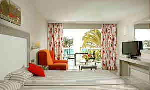 Et hotellrom med to senger, en oransje lenestol, et bord med blomster og en TV. Vinduet viser utsikt over palmer og havet. (AI-generert tekst)