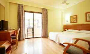 Et lyst hotellrom med to senger, skrivebord, stol og balkong med glassdører. (AI-generert tekst)