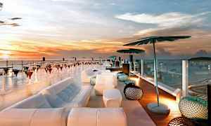 Takbar med utsikt over havet ved solnedgang, med moderne møblering inkludert sofaer, parasoller og bord. (AI-generert tekst)