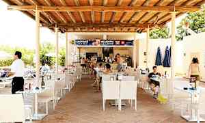 En uteplass ved en bar med bord og stoler under et tretak. Folk sitter ved bordene og bestiller fra baren. (AI-generert tekst)