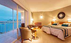 Et hotellrom med to senger, en sittegruppe og utsikt over en balkong med utsikt over byen. (AI-generert tekst)