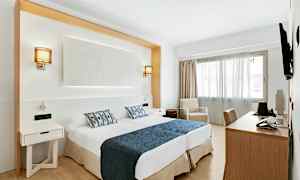 Et hotellrom med to senger med blått sengeteppe og mønstrede puter, skrivebord, stol og en lenestol ved et vindu. (AI-generert tekst)