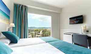 Et hotellrom med to enkeltsenger, et maleri med strandmotiv på veggen, store vinduer med utsikt over landskapet, og en TV på veggen. (AI-generert tekst)