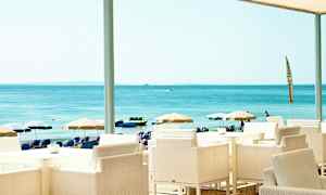 Utsikt fra en strandrestaurant med hvite stoler og bord, i bakgrunnen sees parasoller og havet. (AI-generert tekst)