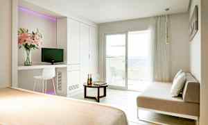 Et lyst hotellrom med en seng, en sofa, et bord og en balkong vendt mot havet. (AI-generert tekst)